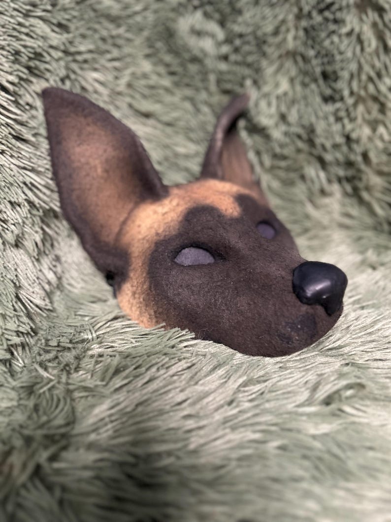 May include: A sculpted dog mask featuring a tan and black coat pattern with large upright ears, a dark muzzle, and a smooth black nose. The mask has deep-set eye sockets and a textured surface finish, capturing the likeness of a Belgian Malinois.