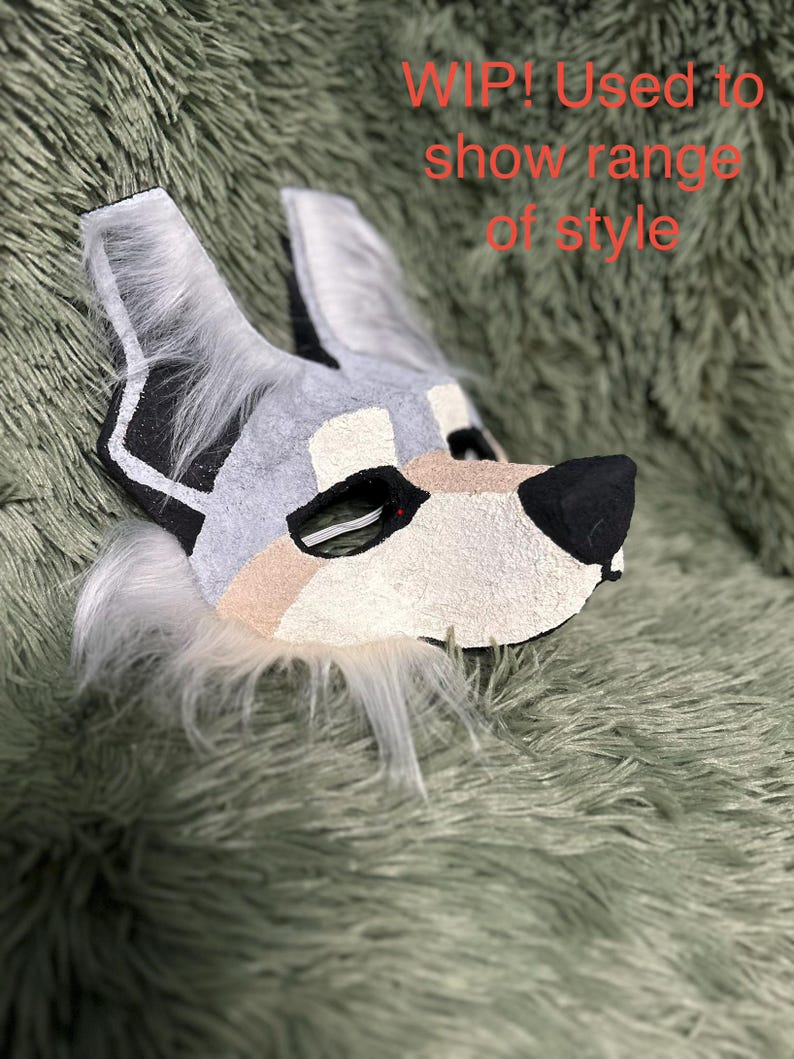 May include: A wolf-like animal mask featuring grey, white, and tan textured panels with black accents around the eyes and nose, and white faux fur trim. The text "WIP! Used to show range of style" is visible in red on the upper right side.