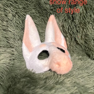 May include: A white and peach-colored animal mask with tall pointed ears and a snout, featuring black-lined eye openings. The text 'WIP! Used to show range of style' is printed in red across the upper portion of the image.