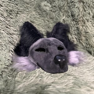 May include: A dark gray animal mask featuring pointed black ears, a smooth black nose, and light purple fur accents along the cheeks. The mask has almond-shaped eye openings and a textured, soft-looking surface covering the entire face structure.
