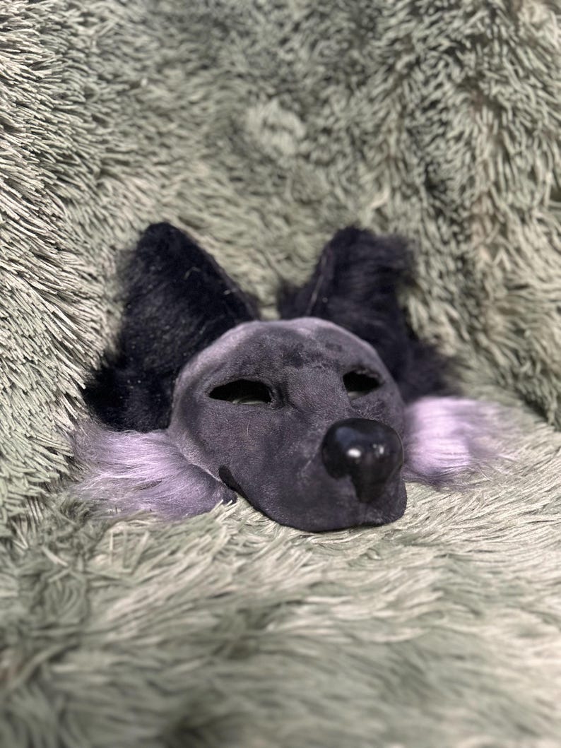 May include: A dark gray animal mask featuring pointed black ears, a smooth black nose, and light purple fur accents along the cheeks. The mask has almond-shaped eye openings and a textured, soft-looking surface covering the entire face structure.