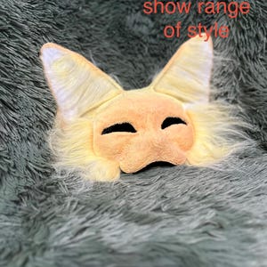 May include: A light yellow and cream-colored animal mask with pointed ears and long faux fur trim. The mask features two dark, almond-shaped eye openings. The text 'WIP! Used to show range of style' is printed in red over the upper portion of the mask.