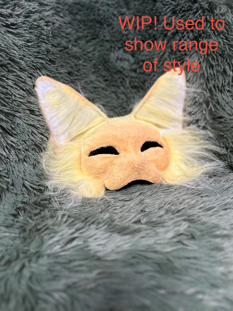 May include: A light yellow and cream-colored animal mask with pointed ears and long faux fur trim. The mask features two dark, almond-shaped eye openings. The text 'WIP! Used to show range of style' is printed in red over the upper portion of the mask.