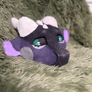 May include: A handcrafted dragon mask featuring a dark purple and black base with light gray horns and accents. The mask has large, expressive teal eyes with black pupils and bright purple ear details, showcasing a textured, felt-like material finish.