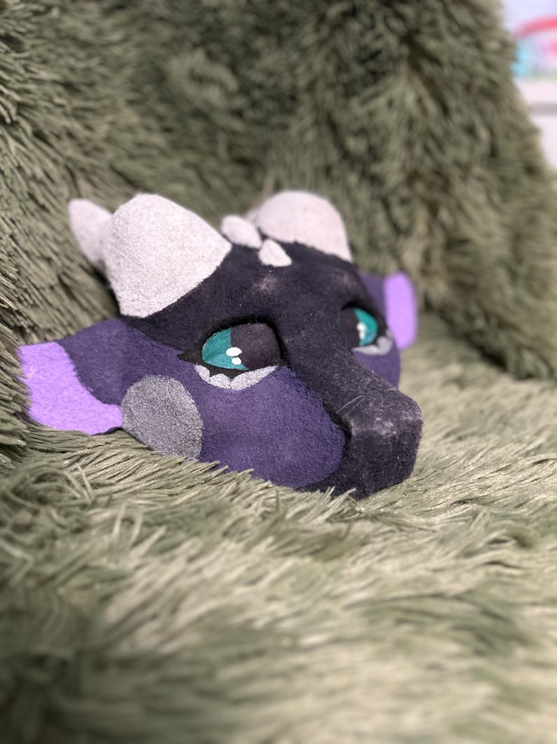 May include: A handcrafted dragon mask featuring a dark purple and black base with light gray horns and accents. The mask has large, expressive teal eyes with black pupils and bright purple ear details, showcasing a textured, felt-like material finish.