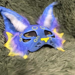 May include: A blue and purple creature mask featuring large pointed ears with white fur accents, yellow fin-like side details, and yellow circular markings around the eyes and nose. The mask has black eye cutouts and a yellow nose patch.