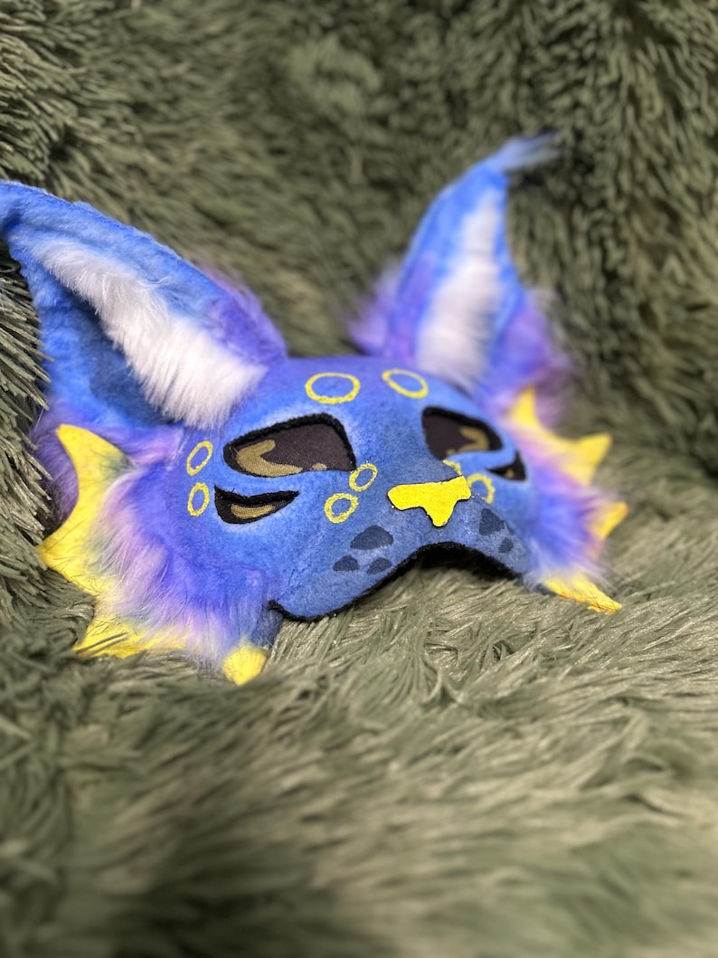 May include: A blue and purple creature mask featuring large pointed ears with white fur accents, yellow fin-like side details, and yellow circular markings around the eyes and nose. The mask has black eye cutouts and a yellow nose patch.