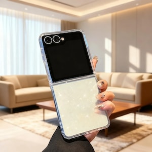 May include: A foldable smartphone encased in a clear protective cover. The upper section features a black screen with two circular camera lenses, while the lower section displays a shimmering, light-colored glitter finish with a sparkling effect.
