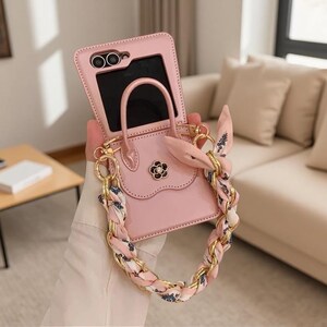 May include: A light pink phone case designed like a miniature handbag with a top handle and a flap closure. The case features a gold-tone chain strap interwoven with a pink and white patterned fabric scarf and a small black and gold flower emblem.