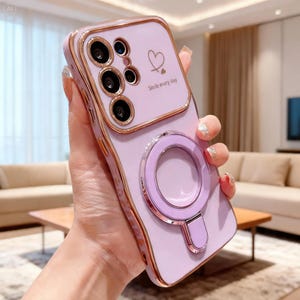 Puede incluir: Una funda para smartphone de color morado claro con un marco dorado metálico alrededor del módulo de cámara y los bordes. La parte trasera presenta un icono de corazón impreso, el texto "Smile every day" y un anillo magnético.