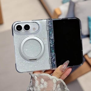 May include: A foldable smartphone case featuring a white glittery back panel with a circular silver magnetic ring mount. The hinge area is decorated with iridescent blue and silver flakes, and the camera cutout accommodates two circular lenses.