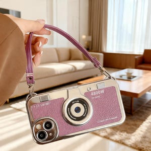 Puede incluir: Una funda de teléfono rectangular diseñada como una cámara vintage, con una superficie texturizada de color rosa malva, un detalle de lente circular central y detalles metálicos plateados, que incluye una correa de muñeca a juego.