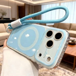Può includere: Una custodia per smartphone azzurro chiaro con bordo trasparente e un anello di ricarica magnetico circolare sul retro. La custodia presenta un ritaglio per tripla fotocamera con bordi argentati e un cinturino da polso intrecciato azzurro.