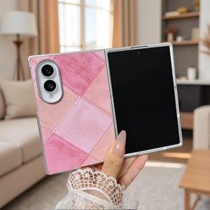 Pode incluir: Uma capa de smartphone dobrável com um design de patchwork de quadrados de camurça em tons de rosa e rosa claro. A capa tem uma moldura de câmara prateada com duas lentes circulares e uma área de ecrã preta no lado direito do dispositivo.