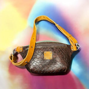 Könnte beinhalten: Eine braune Leder-Bauchtasche mit einem strukturierten Schlangenhautmuster. Die Tasche hat eine Reißverschlusstasche auf der Vorderseite und einen braunen Lederriemen mit einer silbernen Schnalle. Die Tasche hat ein "TRAMWILD" Logo auf der Vorderseite.