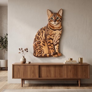 Op de afbeelding: Een houten wandsculptuur van een zittende kat, opgebouwd uit gelaagde houten elementen in diverse bruin- en tinten met donkerbruine vlekken en strepen, spitse oren, groene ogen en een staart die om de voorpoten is gekruld.