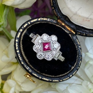 May include: A silver-toned ring featuring a central square-cut red gemstone surrounded by a cluster of round white stones. The band includes baguette-cut stones on each side, all set within a scalloped metal frame resting in a black velvet box.