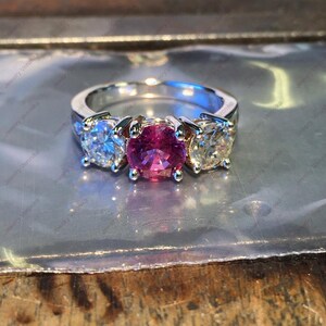 May include: A three-stone ring featuring a central round-cut vibrant pink gemstone flanked by two clear round-cut white gemstones. The stones are set in a polished silver-toned metal band, creating a classic design with a prominent colour contrast.