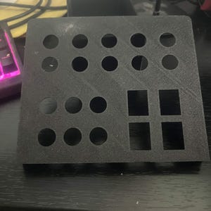 May include: A black, rectangular, 3D-printed plastic panel featuring a grid of ten circular cutouts arranged in two rows at the top, four circular cutouts on the bottom left, and four rectangular cutouts grouped on the bottom right side.