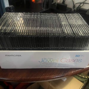 May include: A white cardboard box containing a 50-pack of slim clear jewel cases for CDs or DVDs. The box features the Memorex brand name and the text Slim Clear Jewel Cases 50PK printed on the front in blue and black lettering.