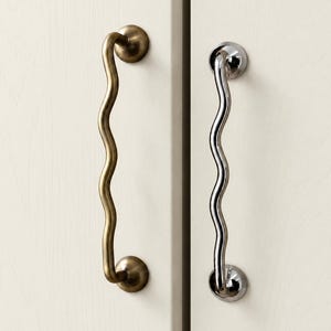 May include: Two vertical cabinet handles with a wavy, serpentine shape. One handle features a matte brass finish, while the other has a polished chrome finish. Each handle is mounted with two circular bases attached to a light-colored cabinet door.