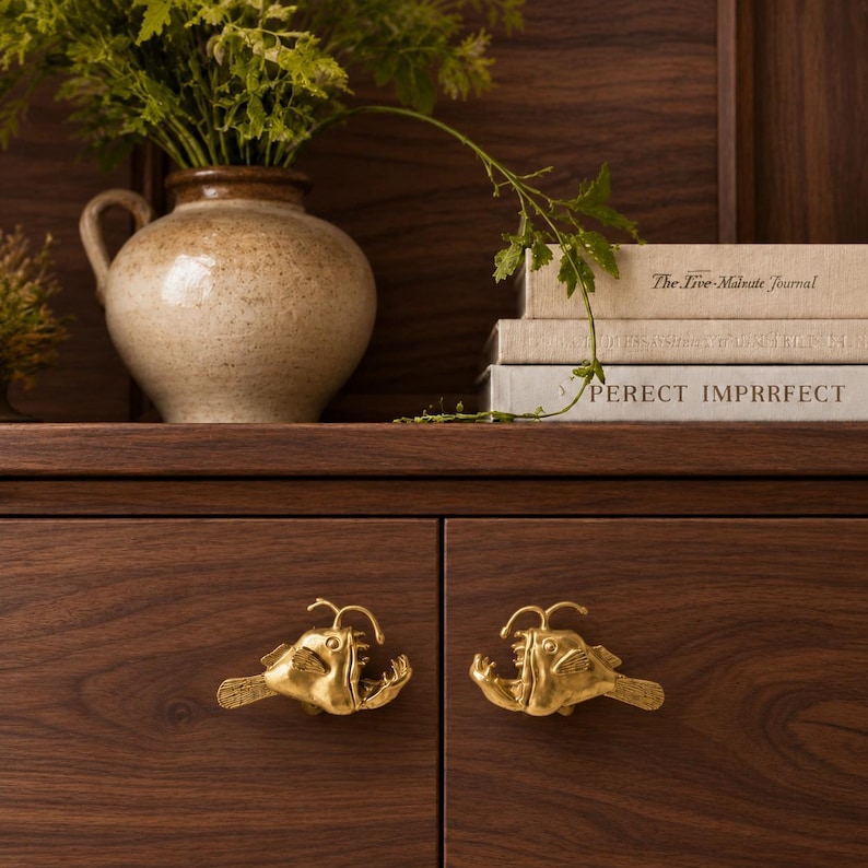 May include: Two gold-coloured metal cabinet knobs shaped like anglerfish with detailed fins and a lure. The knobs are mounted on dark brown wooden cabinet doors. The fish figures feature textured scales and open mouths with visible teeth.