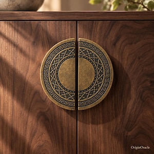 May include: A pair of semi-circular cabinet pulls made of antique brass with a dark, recessed background. Each handle features a detailed Greek key border and a central quatrefoil pattern, mounted on a dark brown wood cabinet door.