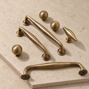 Op de afbeelding: Een collectie messingkleurig kastbeslag bestaande uit twee lange gebogen ladehandgrepen, één kortere gebogen handgreep, twee bolvormige knoppen en één T-vormige knop. Elk onderdeel heeft een gladde, matte metalen afwerking en afgeronde randen.