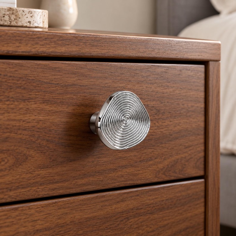 May include: A silver-toned cabinet knob with an irregular, rounded triangular shape and a concentric, rippled texture on its face. The knob is mounted on a dark brown wooden drawer front with a visible wood grain pattern.