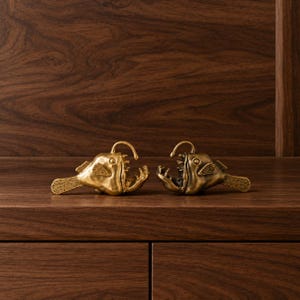 May include: Two metallic brass-coloured anglerfish figurines facing each other. Each fish features a prominent dorsal lure, wide open jaws with visible sharp teeth, textured fins, and a detailed tail. The figures have a polished, reflective finish.