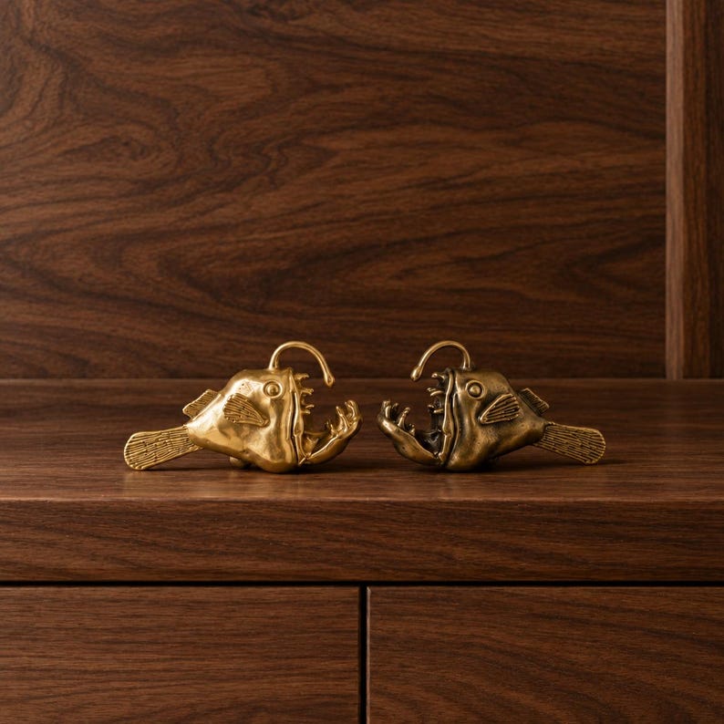 May include: Two metallic brass-coloured anglerfish figurines facing each other. Each fish features a prominent dorsal lure, wide open jaws with visible sharp teeth, textured fins, and a detailed tail. The figures have a polished, reflective finish.