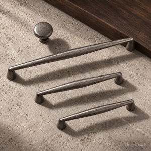 May include: A set of dark gray metal cabinet hardware including one round knob and three elongated drawer pulls of varying lengths. Each piece features a textured, weathered finish with tapered ends and a central raised profile.