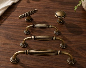 May include: A collection of antique brass cabinet hardware including three arched drawer pulls with ribbed detailing, one T-shaped handle with vertical grooves, and one circular knob with a smooth, slightly domed surface and a matching metallic finish.
