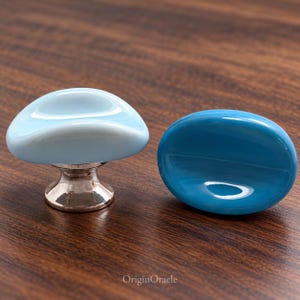 May include: Two cabinet knobs with smooth, rounded, glossy surfaces. One knob is light blue with a metallic silver base, while the other is a solid, darker blue oval shape. The text OriginOracle is visible at the bottom center.