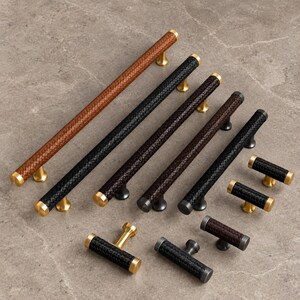 May include: A collection of cabinet pulls and knobs featuring a woven leather-textured surface. The hardware comes in tan, black, and dark brown, accented with metallic gold or dark grey cylindrical end caps and mounting posts.