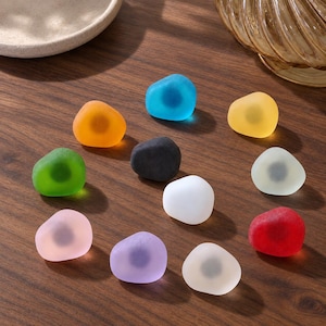 Könnte beinhalten: Eine Sammlung von elf glatten, abgerundeten, mattierten Glassteinen in verschiedenen Farben wie Blau, Orange, Grün, Schwarz, Weiß, Gelb, Cremeweiß, Rosa, Violett und Rot, die jeweils einen dunklen, kreisförmigen Magnetkern besitzen.
