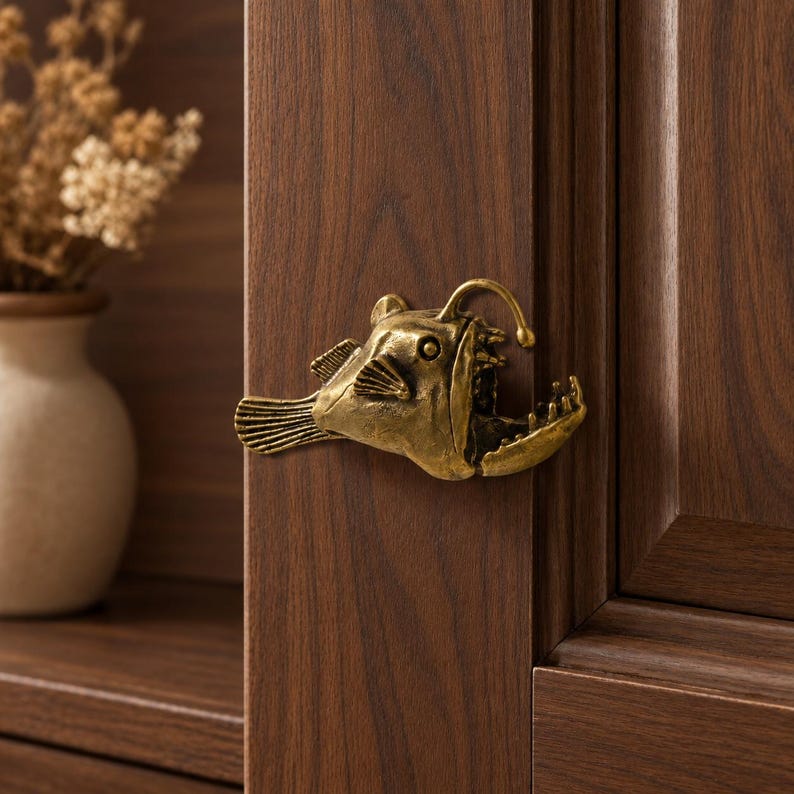 May include: A brass-toned metal cabinet knob shaped like an anglerfish with a prominent lure, fins, and an open mouth with visible teeth, mounted on a dark brown wood-grained cabinet door surface with a vertical panel design.