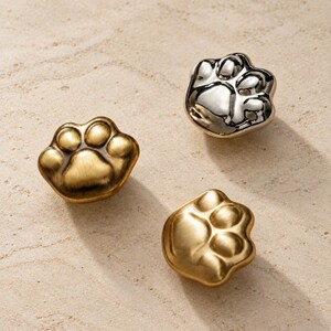 May include: Three metal cabinet knobs shaped like animal paw prints. Two of the knobs have a brushed gold finish, while the third knob features a polished silver finish. Each knob displays a central pad and four distinct toe pads in relief.