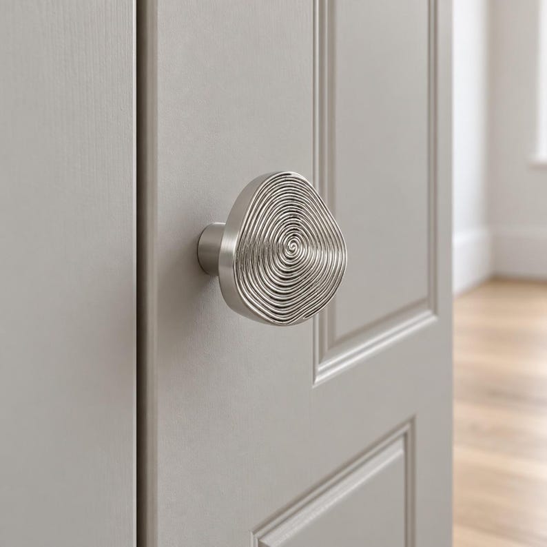May include: A silver-toned cupboard knob with an irregular, rounded shape and a textured surface featuring concentric, swirling circular lines. The knob is mounted on a light grey panelled door, providing a metallic accent to the surface.