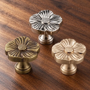 May include: Three metal cabinet knobs shaped like flowers with textured petals and a central stamen detail. One knob has a polished silver finish, one has an antique brass finish, and one has a brushed gold finish, each with a pedestal base.