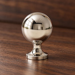 Pode incluir: Um puxador de móvel em metal prateado polido com uma cabeça esférica sobre uma base cilíndrica cónica. A superfície lisa e refletora apresenta reflexos nítidos, criando um aspeto metálico brilhante para ferragens de mobiliário.