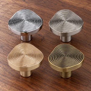 May include: Four cabinet knobs with an irregular, rounded triangular shape. Each knob features a raised, concentric spiral pattern on the top surface. The set includes one polished chrome, one brushed nickel, one brushed brass, and one antique bronze finish.
