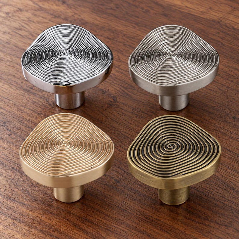 May include: Four cabinet knobs with an irregular, rounded triangular shape. Each knob features a raised, concentric spiral pattern on the top surface. The set includes one polished chrome, one brushed nickel, one brushed brass, and one antique bronze finish.