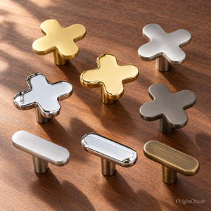 Op de afbeelding: Een collectie kastknoppen in diverse afwerkingen, waaronder geborsteld goud, gepolijst chroom, mat zilver en antiek messing. De set bevat kruisvormige en langwerpige ovale ontwerpen. De tekst OriginOracle is zichtbaar in de hoek.