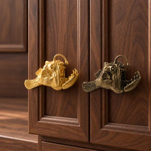 Puede incluir: Dos pomos para armarios con forma de pez rape, uno con acabado dorado brillante y otro con acabado en bronce antiguo. Cada pomo presenta el señuelo característico del pez, aletas y una boca abierta con dientes visibles sobre una puerta.
