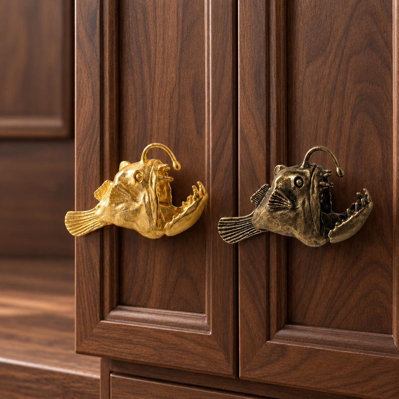 May include: Two cabinet knobs shaped like anglerfish, one with a bright gold finish and the other with an antique bronze finish. Each knob features the fish's characteristic lure, fins, and an open mouth with visible teeth, mounted on a wooden door.