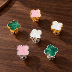 May include: Six clover-shaped cabinet knobs featuring smooth, marbled-effect inserts in pink, green, and white. Three knobs have polished gold-tone bases, while the other three feature polished silver-tone bases, each with a rounded, tiered stem.