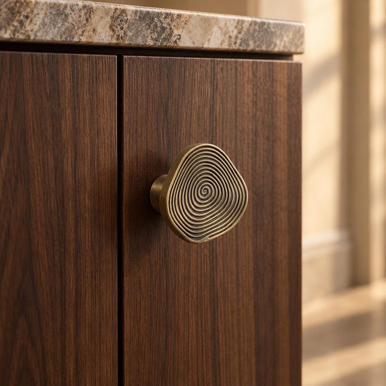 May include: A brass-toned cabinet knob with an irregular, organic shape and a concentric spiral pattern engraved on its front surface, mounted on a dark brown wood-grain cabinet door with a visible vertical grain pattern.
