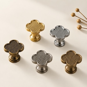 May include: Five clover-shaped cabinet knobs arranged in a group. Each knob features a textured grid pattern on the top surface and a smooth, tapered base. The set includes finishes in polished gold, bright silver, brushed nickel, and antique brass.
