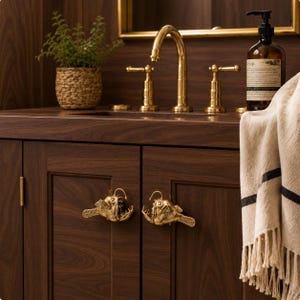 May include: Two brass cabinet knobs shaped like anglerfish with detailed fins and open mouths, mounted on dark wood cabinet doors. The knobs feature a metallic gold finish and are positioned horizontally on the front of the vanity doors.